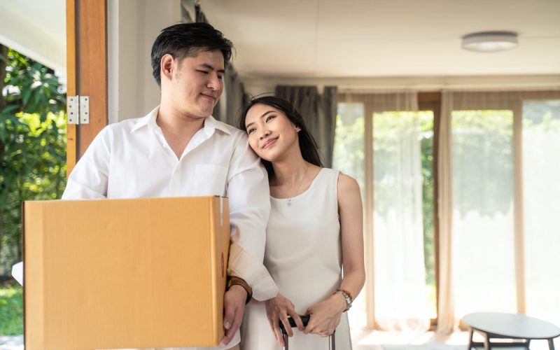 Asian couple moving to new house, man holding big box standing near pregnancy woman